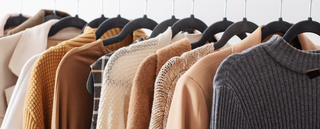 Zdjęcie przedstawia ubrania, między innymi swetry w jesiennych kolorach zawieszone na czarnych wieszakach.